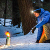 Wearing a headlamp and blue jacket, someone kneels on snowy ground in a forest, using the American Reserves Kelly Kettle® Ultimate Trekker Camping Kit to heat water on their portable camping stove.