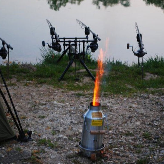 A metal stove showcases a visible flame on the riverbank beside an American Reserves Kelly Kettle® Ultimate Trekker Camping Kit, with fishing rods on stands by the waters edge.