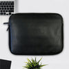The PU Leather Large Privacy Protection Laptop Sleeve (9 x 13.5) by American Reserves lies on a white surface next to a partially visible laptop, small plant, and paper.
