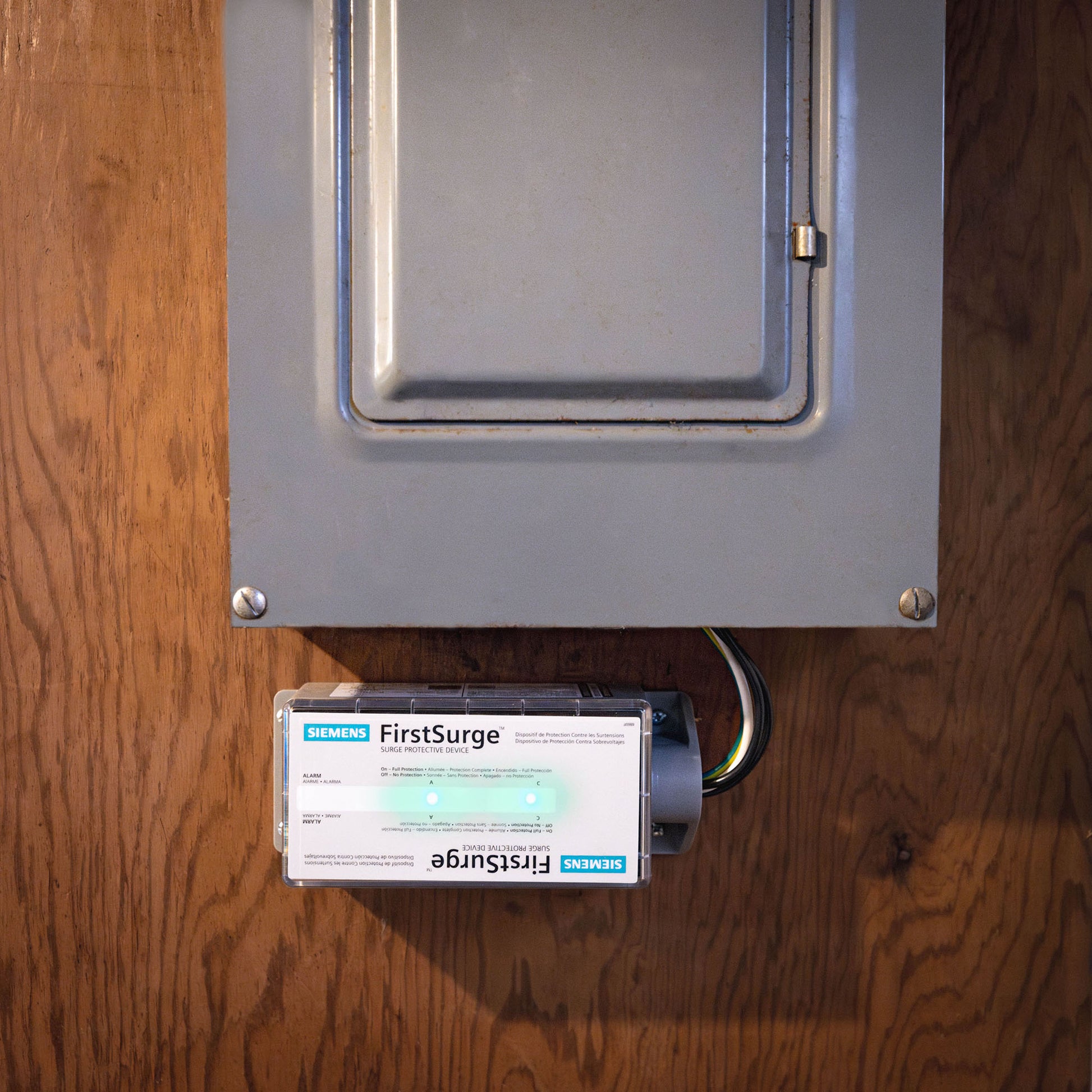 An American Reserves SIEMENS FS™ PRO 140 + Whole House Ferrites EMP Home Protection Kit, noted for its surge protection abilities, is installed below a closed gray electrical panel on a wooden wall.