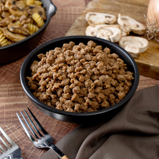 A bowl of cooked beef from the American Reserves 110 Serving Freeze Dried Meat & Sides Bucket sits on a wooden table, surrounded by uncooked spiral pasta and sliced mushrooms on a cutting board. Ideal for emergencies, it promises quality nourishment. Nearby are a napkin and two forks.