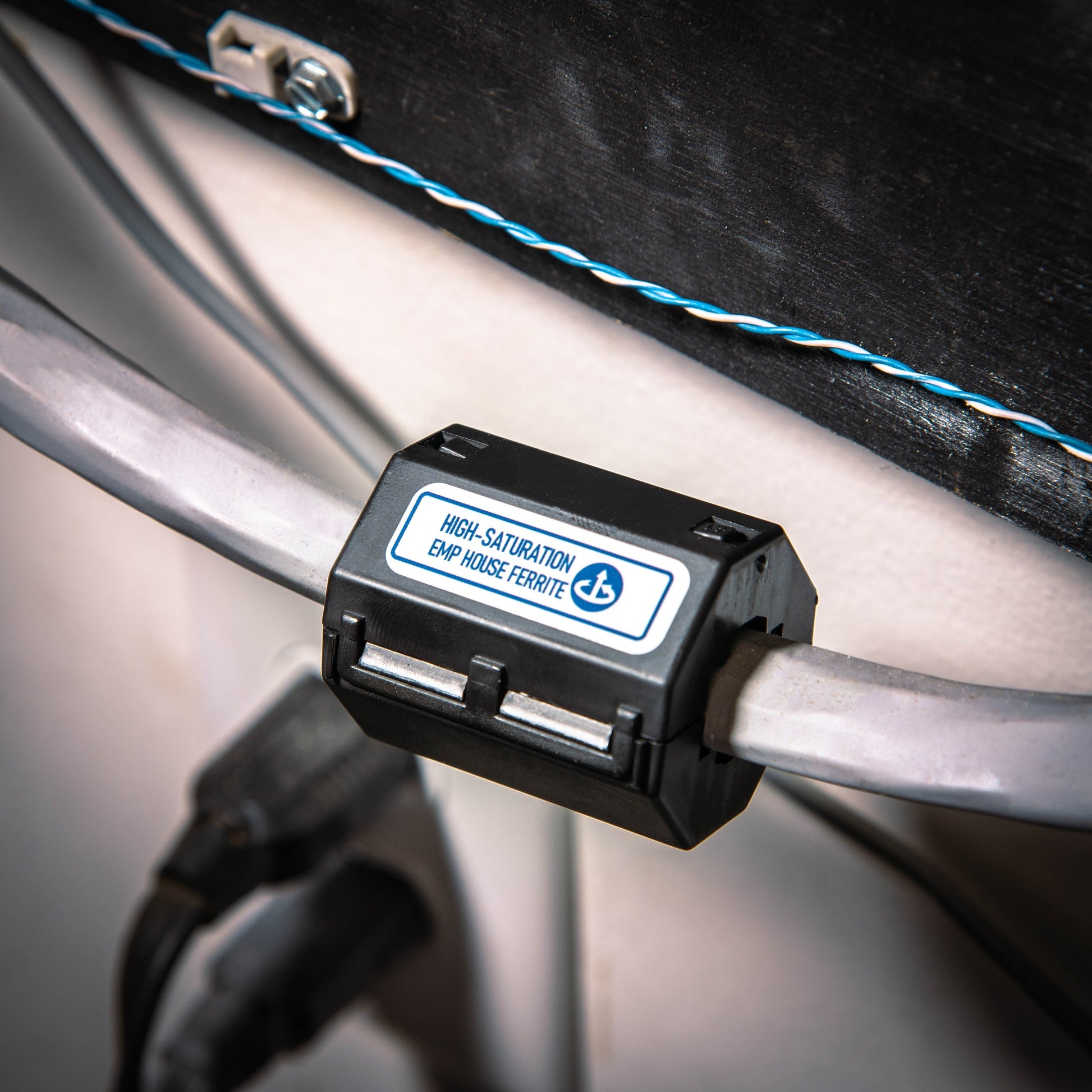 A high-saturation snap ferrite, part of the Complete Auto And Home Protection EMP Kit by American Reserves, is clamped onto a cable. In the background, theres a black panel and a twisted blue-white wire.