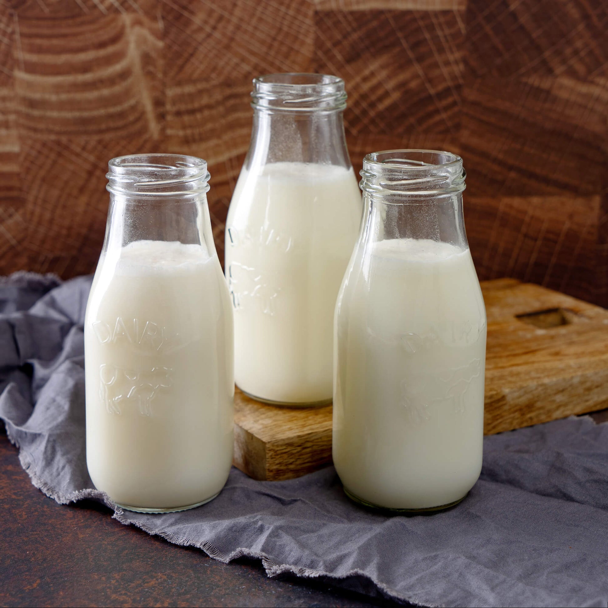 Three glass milk bottles on a wooden board and crumpled gray cloth stand against a rich brown texture. Perfect for emergencies, these bottles promise freshness and reliability alongside your American Reserves 30 Day Emergency Food Supply.